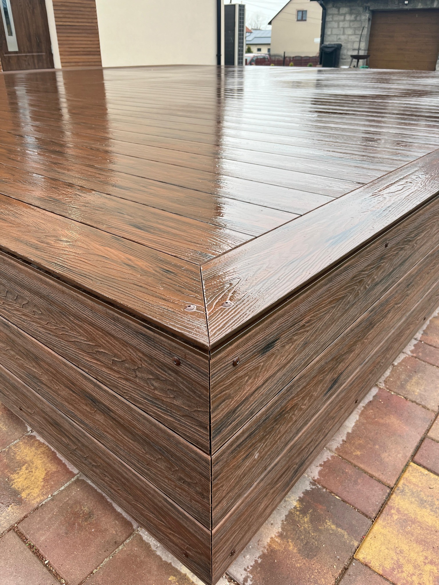 Mokry, brązowy taras z desek kompozytowych, z widocznym detalem narożnika i fakturą drewna, po deszczu, na tle kostki brukowej.