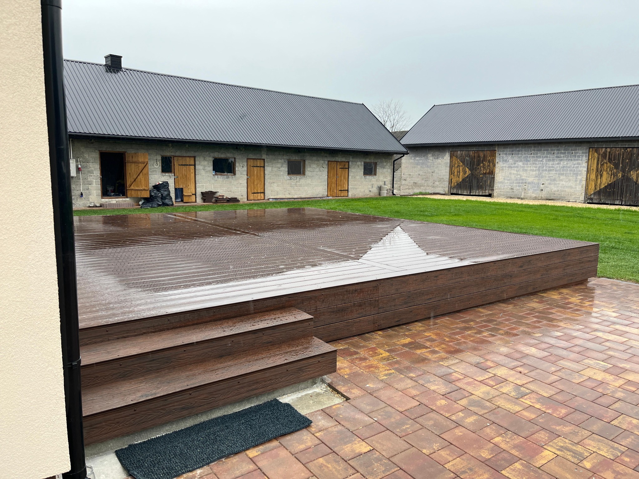 Deszczowy dzień na tarasie z kompozytu. Widoczne schody, deski i budynki gospodarcze w tle. Ceglana kostka brukowa. Matowa wycieraczka przy schodach.