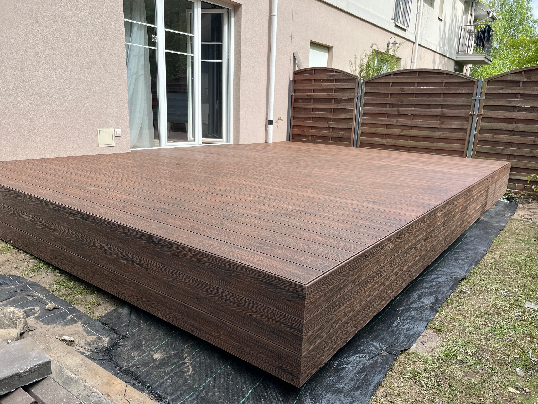 Nowoczesny taras w kolorze ciemnego drewna, z widoczną strukturą desek, tuż przy budynku z dużymi oknami i ogrodzeniem w tle. Widoczna czarna mata pod tarasem.