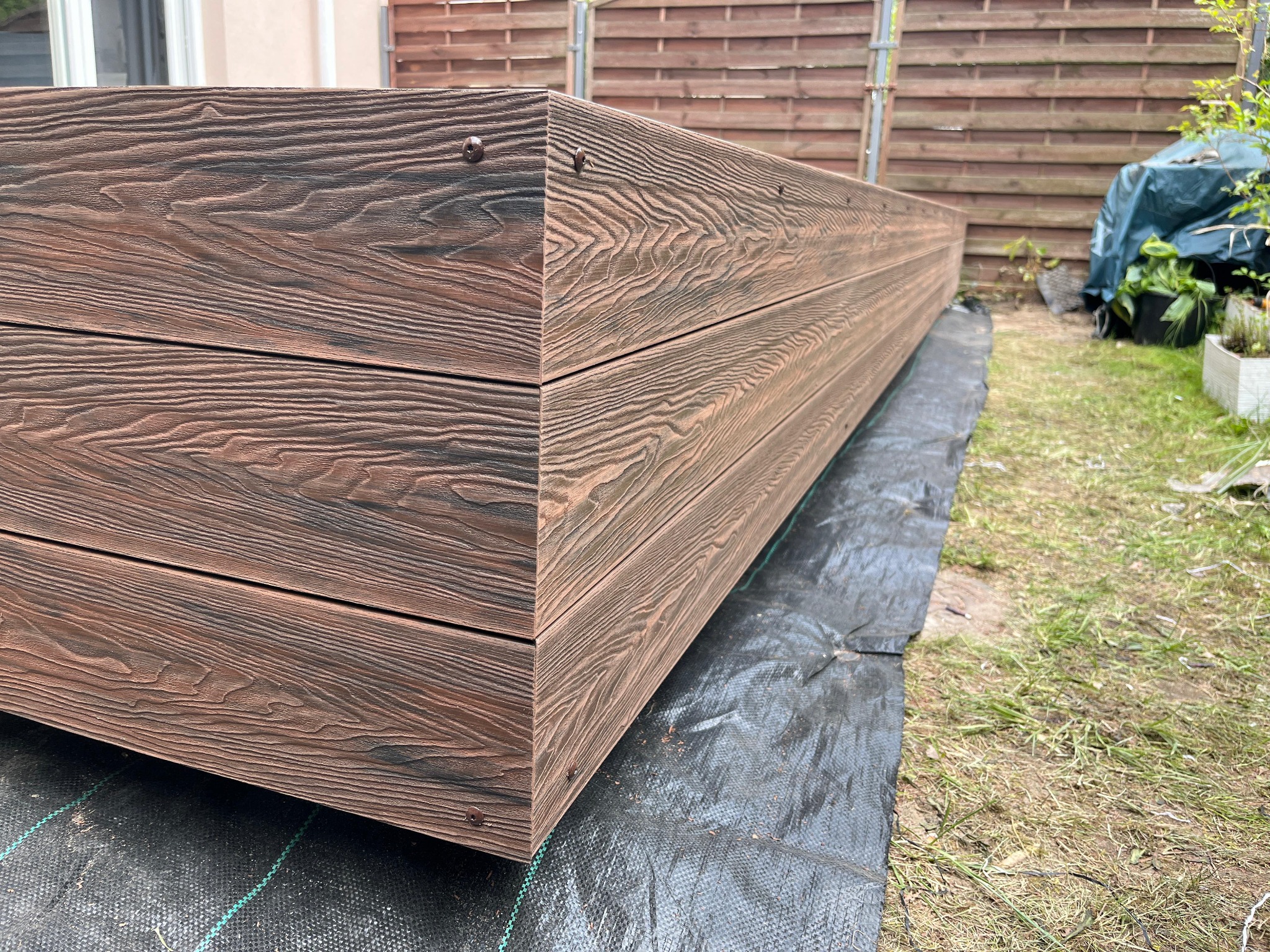 Krawędź podwyższonej rabaty z desek kompozytowych o wyraźnym rysunku drewna, mocowanych na wkręty. Widoczny fragment ogrodu i brązowe, drewniane ogrodzenie.