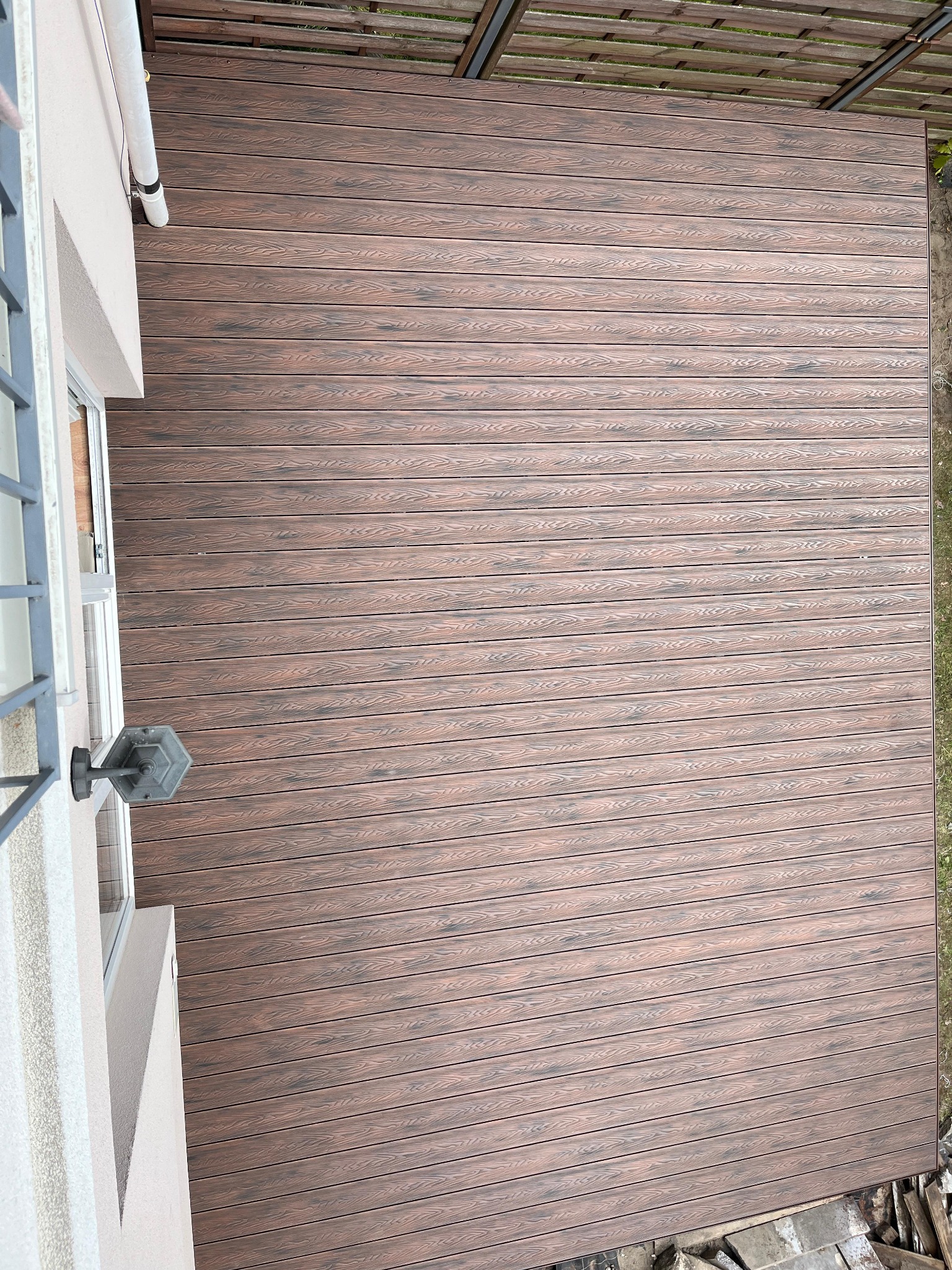 Kompozytowy taras w odcieniach brązu, ułożony przy ścianie budynku z oknem i lampą. Widok z góry, ukazujący strukturę desek i estetyczne wykończenie.