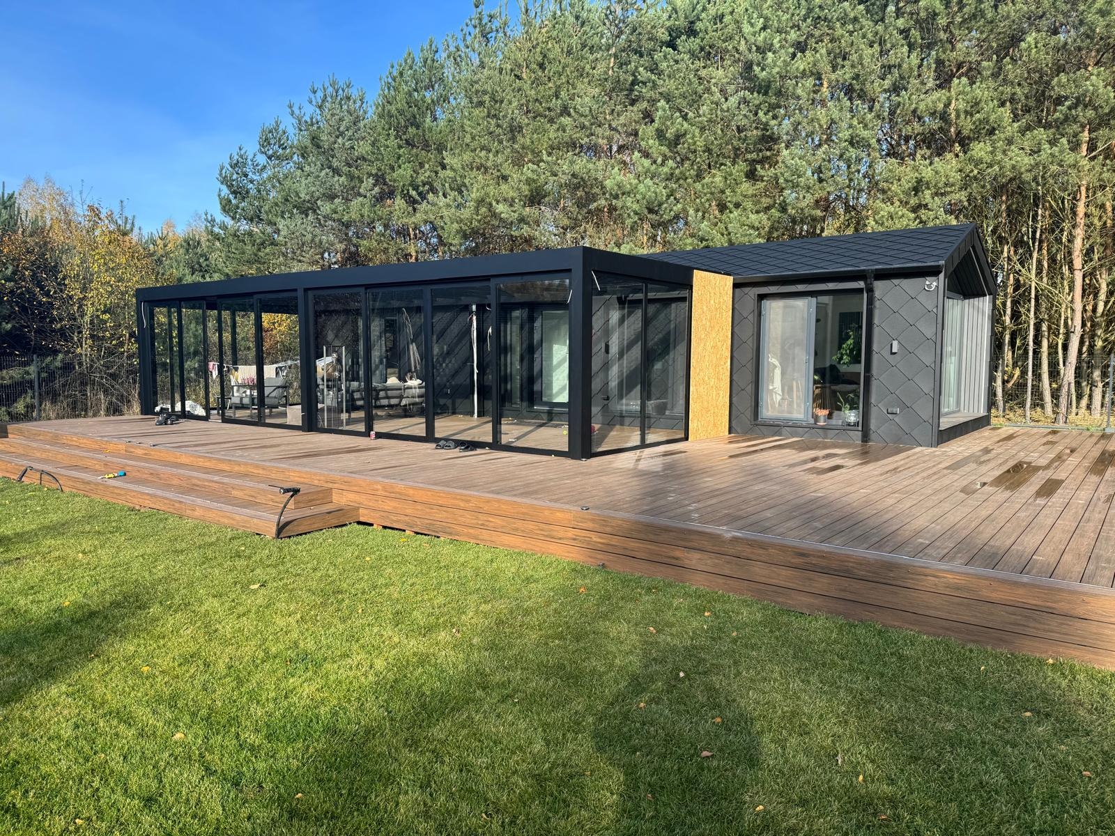 Nowoczesna, przeszklona altana z czarną ramą, połączona z małym domkiem na drewnianym tarasie. Otoczenie: trawnik i drzewa. Widok z poziomu trawnika.