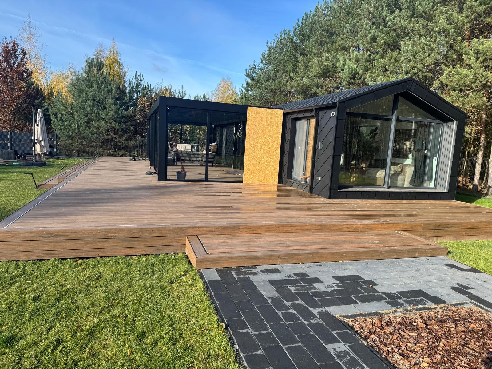 Nowoczesny dom z dużym tarasem z desek i przeszkloną zabudową. Widoczne elementy wykończeniowe i fragment ścieżki z kostki brukowej. Otoczenie: trawa i drzewa.