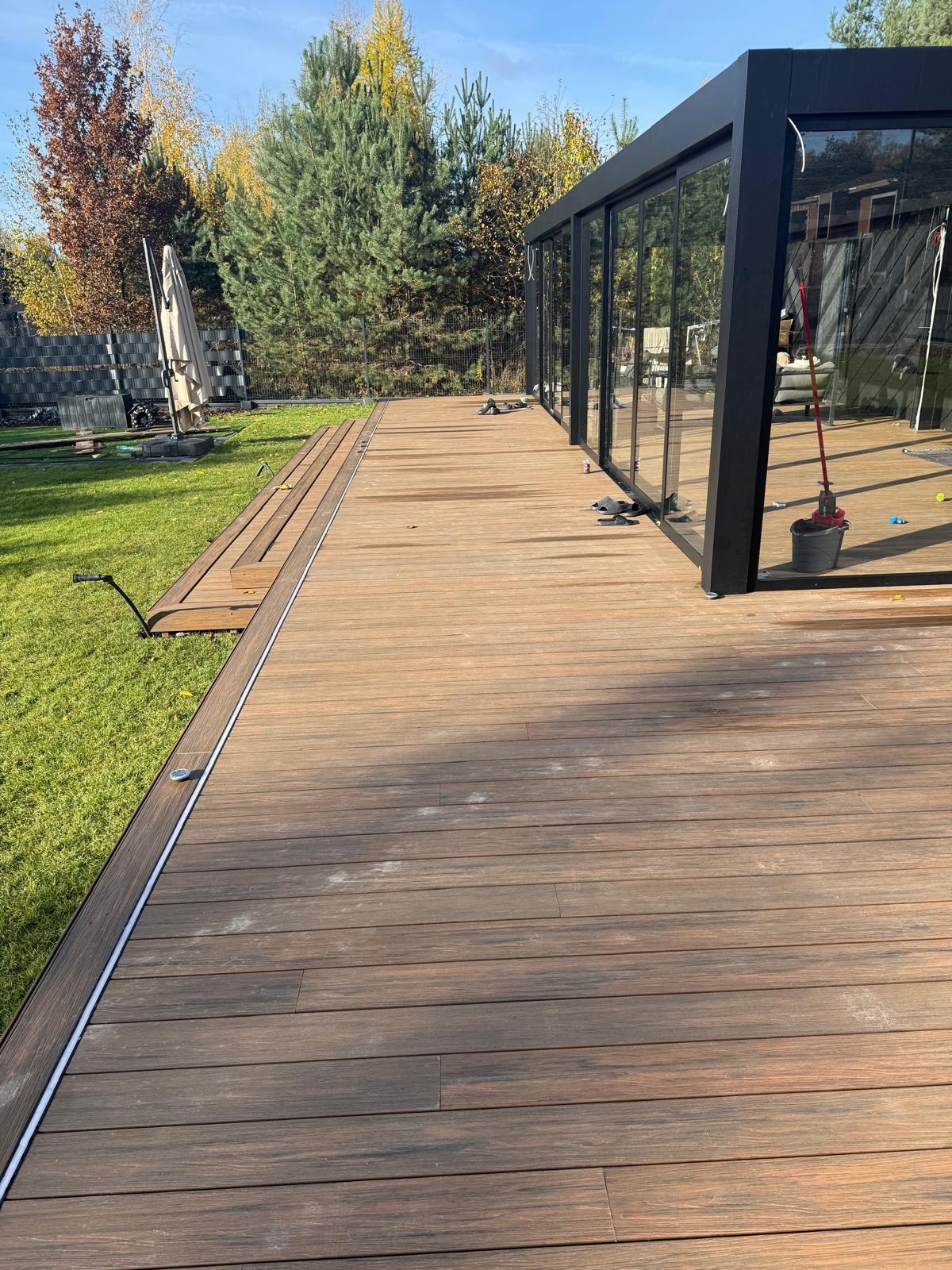 Nowoczesny taras kompozytowy z ciemnobrązowych desek, przylegający do przeszklonej konstrukcji. Widoczne detale wykończenia i zieleń trawnika w tle.
