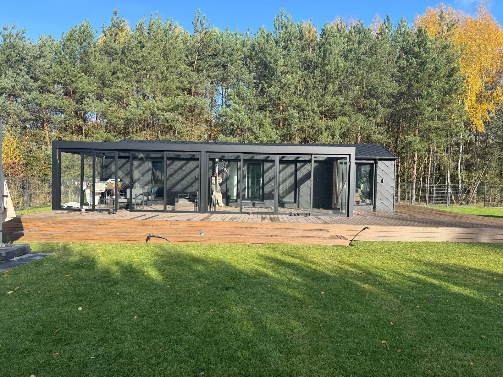 Nowoczesna, przeszklona altana z czarną ramą na drewnianym tarasie, otoczona zielonym trawnikiem i drzewami. Widok na wnętrze z meblami.