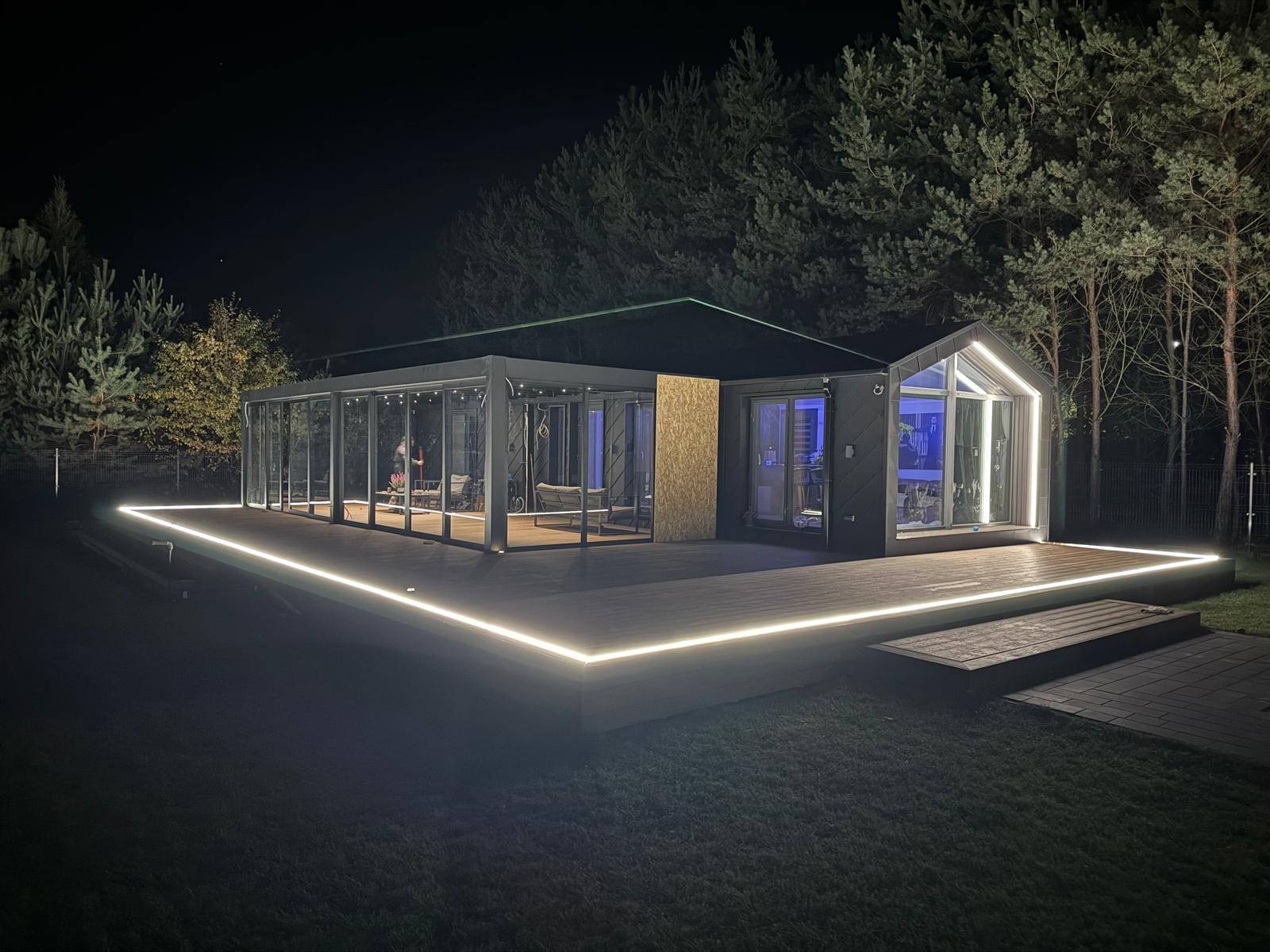 Nowoczesna zabudowa tarasowa z oświetleniem LED w Radomiu. Szklana konstrukcja połączona z drewnianym tarasem, oświetlona nocą, otoczona zielenią. Unikalny projekt.