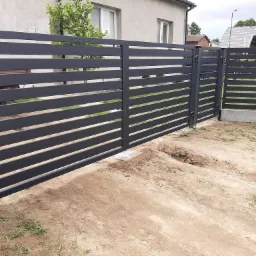 Nowoczesna, pozioma brama wjazdowa z szarych, metalowych profili, częściowo otwarta, z widocznymi śladami prac ziemnych w tle domu.