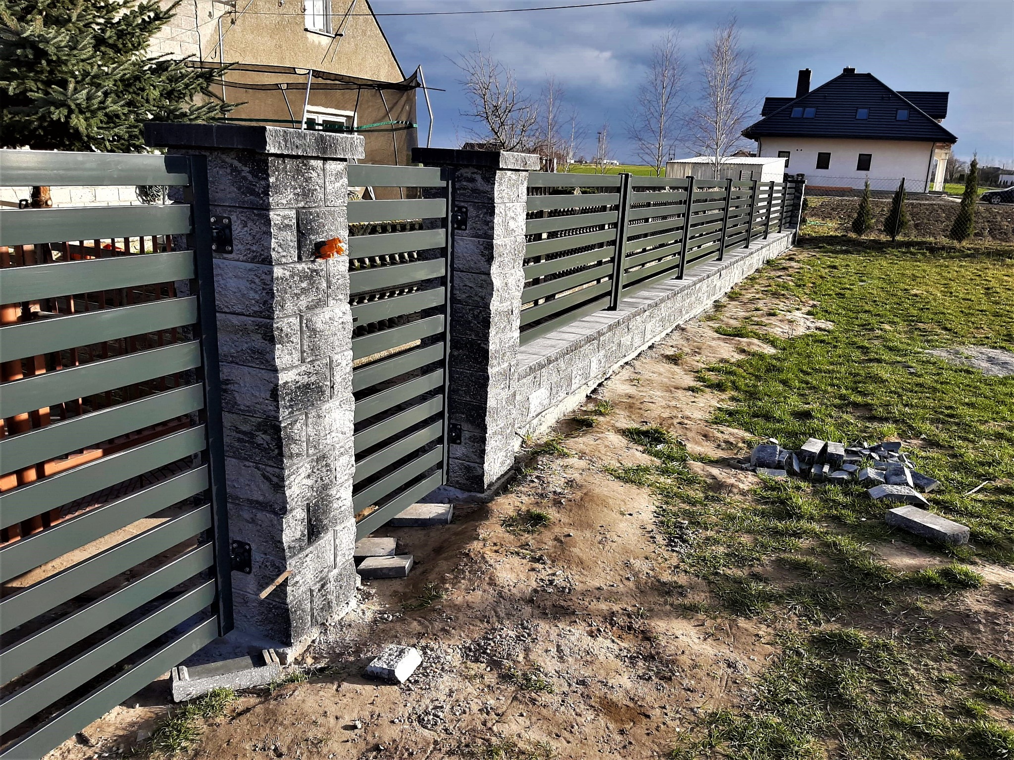 Nowoczesne ogrodzenie panelowe z poziomych, szarych listew, osadzone na słupkach z szarej kostki łupanej, z otwartą bramą wjazdową i widocznymi niedokończonymi pracami wykończeniowymi wokół, na tle...