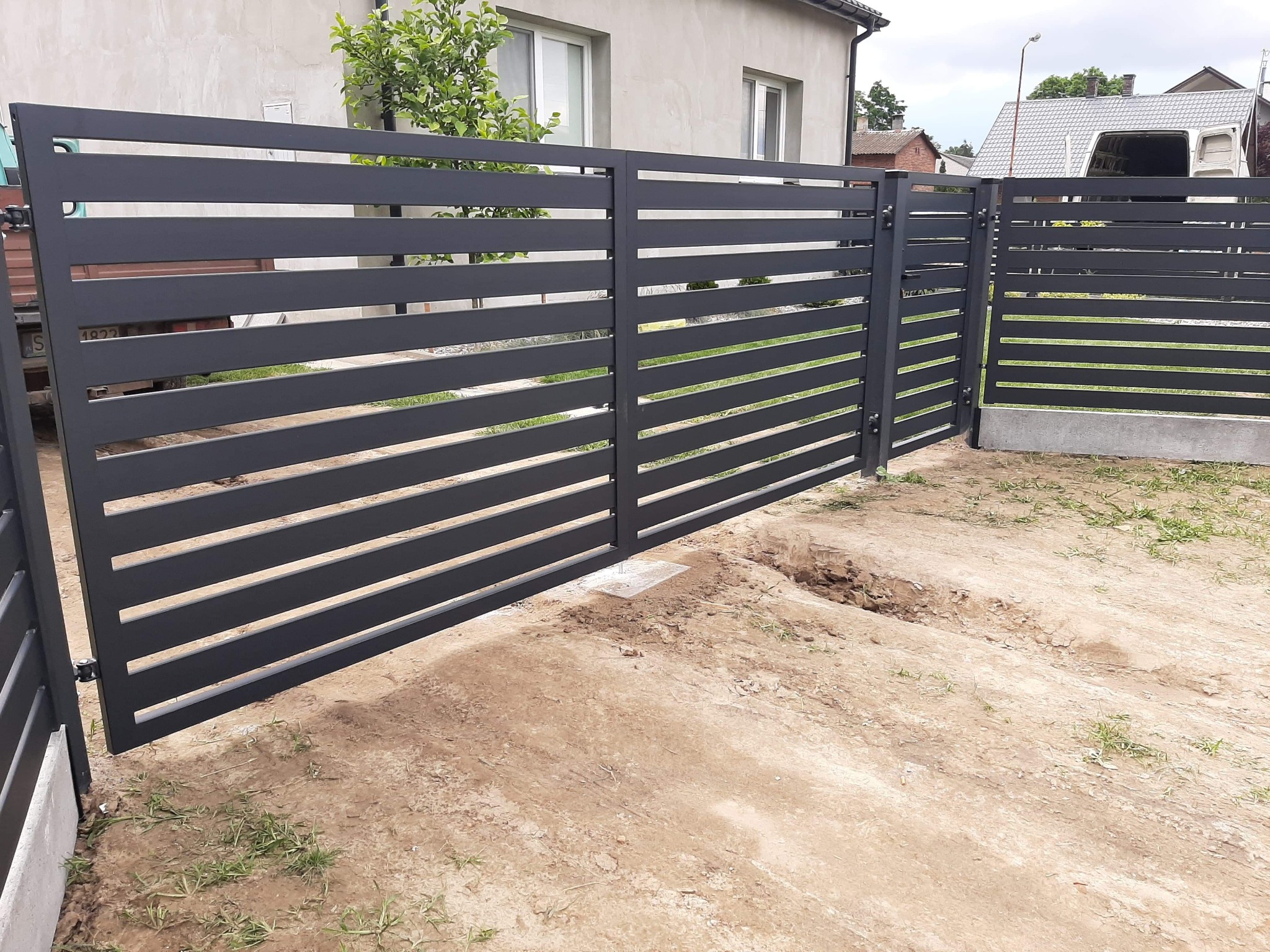 Nowoczesna, pozioma brama wjazdowa z szarych, metalowych profili, częściowo otwarta, z widocznymi śladami prac ziemnych w tle domu.