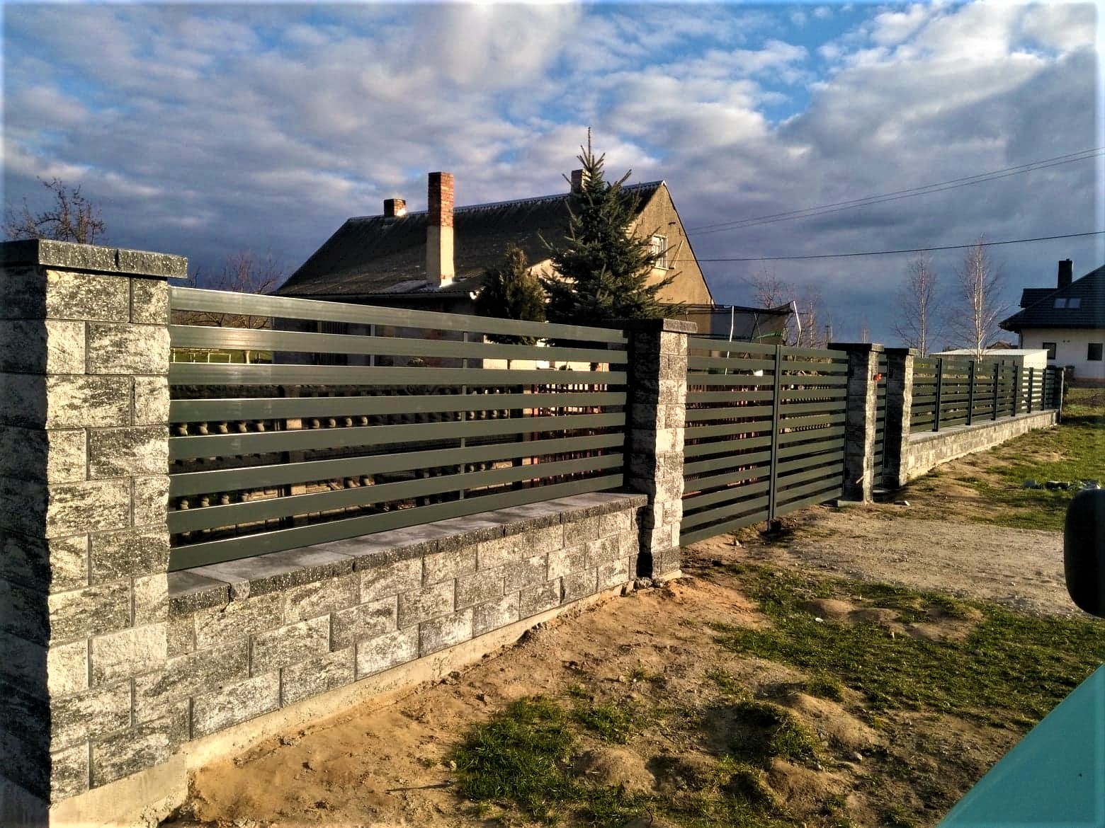 Nowoczesne ogrodzenie z poziomych, metalowych profili w kolorze grafitowym, osadzone na podmurówce i słupkach z szarej kostki łupanej, na tle domu z ogrodem pod błękitnym, zachmurzonym niebem.
