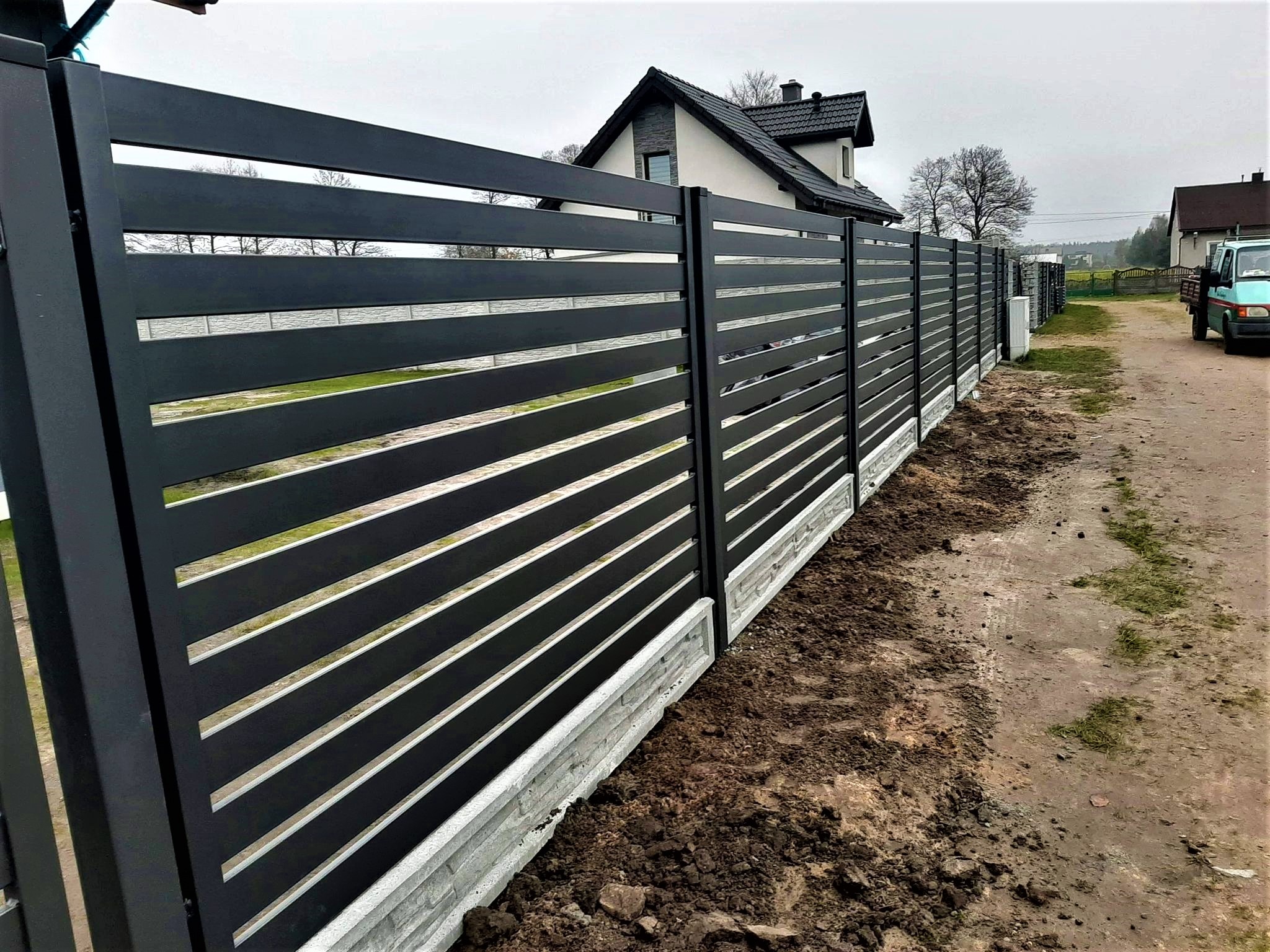 Nowoczesne, poziome przęsła w kolorze grafitowym, zamontowane na betonowej podmurówce, tworzące ogrodzenie posesji z widokiem na dom i zielone otoczenie w pochmurny dzień.