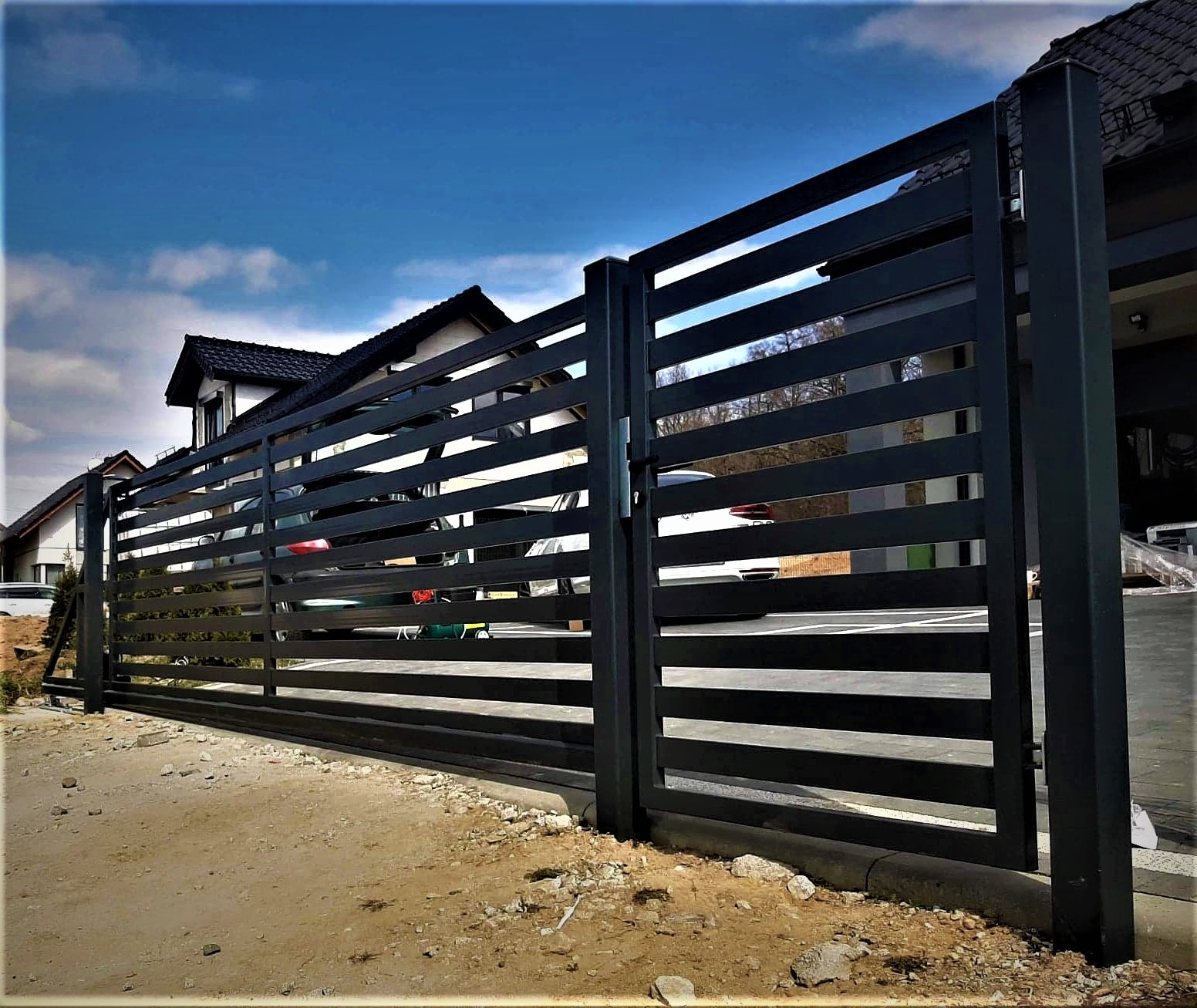 Nowoczesna, pozioma brama wjazdowa z metalowych profili, pomalowana na czarno, na tle domu z ciemnym dachem i błękitnego nieba.