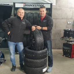 Dwóch mężczyzn w warsztacie w czarnych kurtkach marki Bridgestone pozuje obok stosu opon zimowych i maszyn do montażu kół.