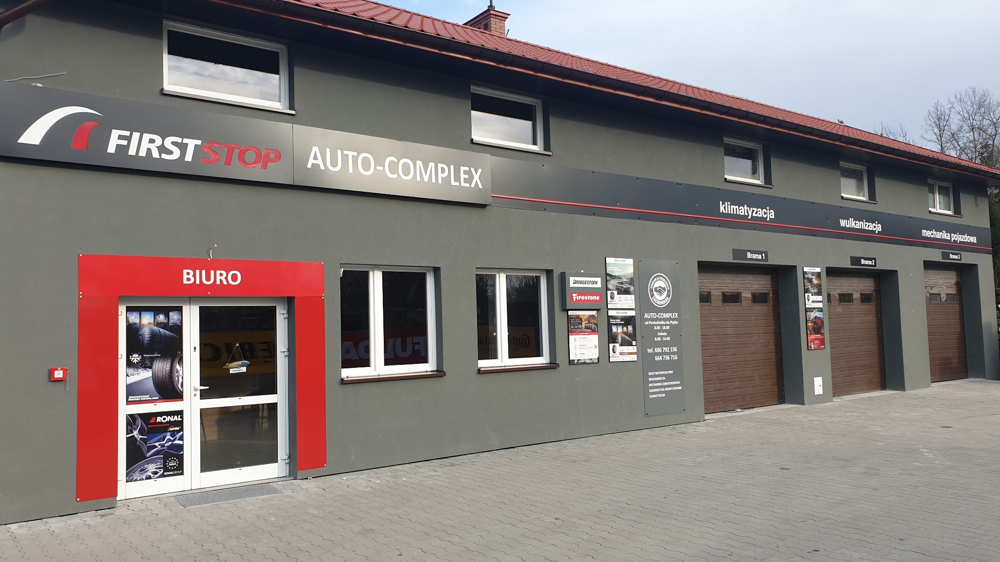 Fasada budynku warsztatu samochodowego First Stop Auto-Complex z trzema bramami garażowymi, wejściem do biura z plakatami reklamowymi opon i felg, oraz tablicami informacyjnymi dotyczącymi...