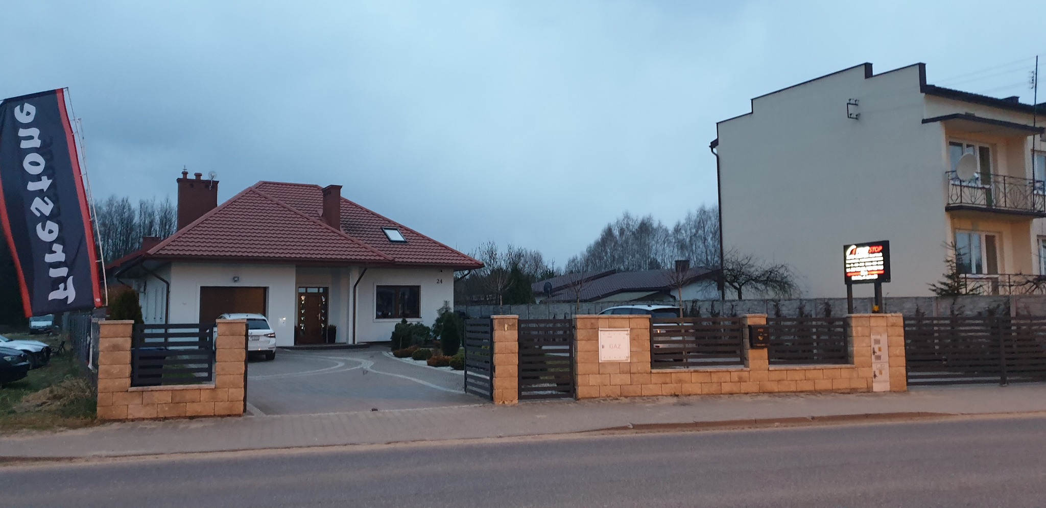 Budynek warsztatu samochodowego z podjazdem, zaparkowanym samochodem i reklamą świetlną przy drodze.