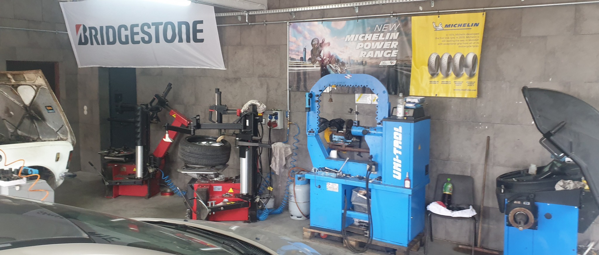 Wnętrze warsztatu samochodowego z dwoma samochodami z otwartymi maskami, urządzeniami do wymiany opon, banerem Bridgestone i plakatami Michelin na ścianie.