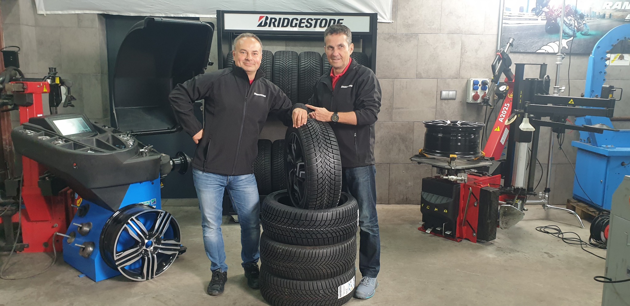 Dwóch mężczyzn w warsztacie w czarnych kurtkach marki Bridgestone pozuje obok stosu opon zimowych i maszyn do montażu kół.