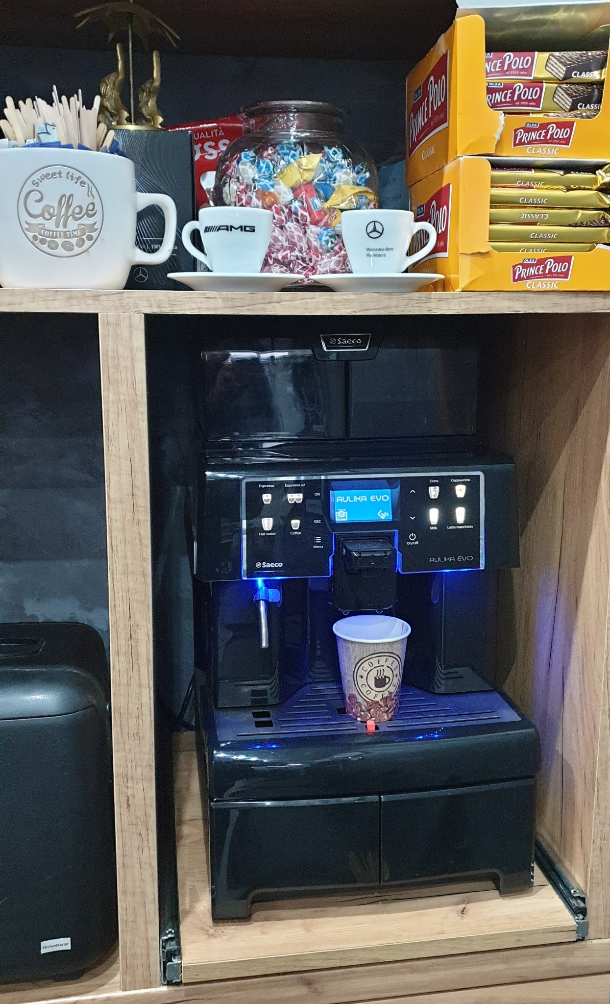 Ekspres do kawy Saeco Aulika EVO w zabudowie meblowej, z filiżanką kawy na podstawce i kolekcją słodyczy oraz filiżanek z logo Mercedesa na półce powyżej.