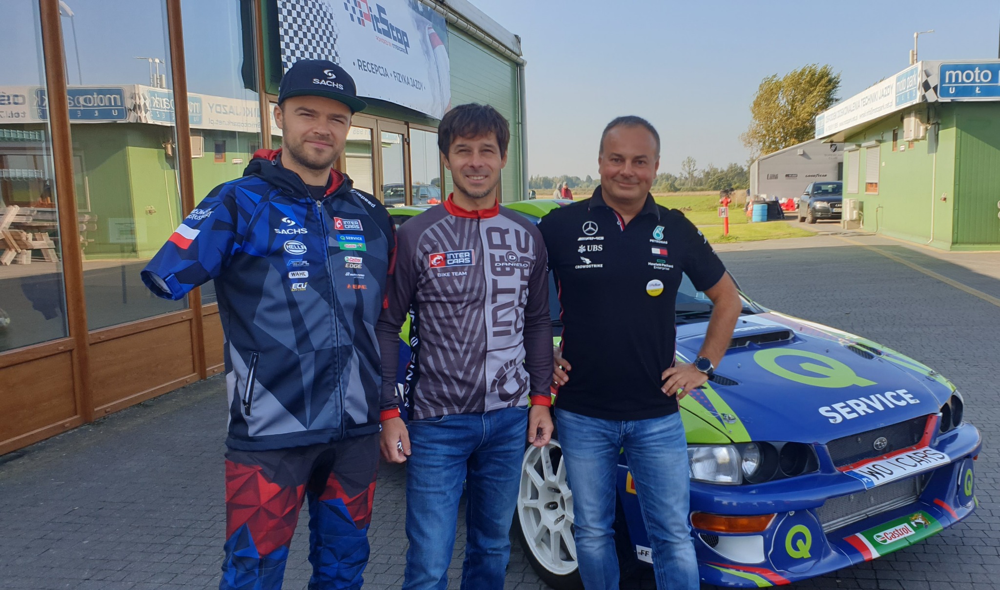 Trzech mężczyzn w odzieży sportowej pozuje na tle niebieskiego samochodu rajdowego Subaru Impreza z zielonymi akcentami i logotypami sponsorów, budynek z napisem 'Recepcja Fizyka Jazdy' w tle.