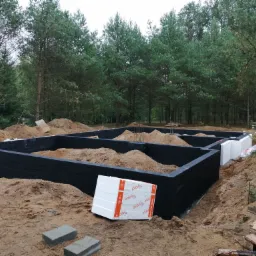 Budowa fundamentów domu murowanego: czarne ściany fundamentowe, częściowo zasypane piaskiem, z widoczną izolacją termiczną i stertą styropianu, na tle leśnego krajobrazu.