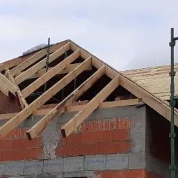 Wznoszenie drewnianej konstrukcji dachu na murowanym domu z czerwonej cegły, widoczne rusztowanie i częściowo pokryte deskami poszycie.