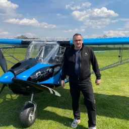 Mężczyzna w czarnym dresie stoi przed małym, niebiesko-czarnym samolotem Bush Cat na trawiastym lotnisku w słoneczny dzień.