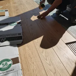 Montaż jasnych, dębowych desek podłogowych z użyciem gumowego młotka, widoczne kartony producenta Finishparkiet z logo 50 lat gwarancji.