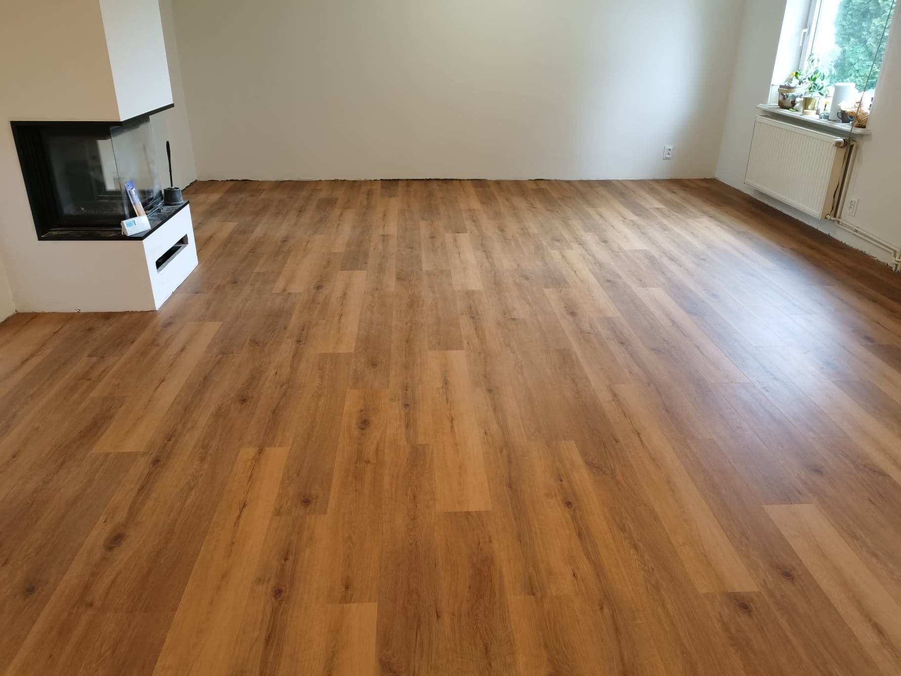 Ukończona instalacja paneli podłogowych w pustym pokoju z kominkiem i grzejnikiem pod oknem, widoczne niedociągnięcia przy ścianach.