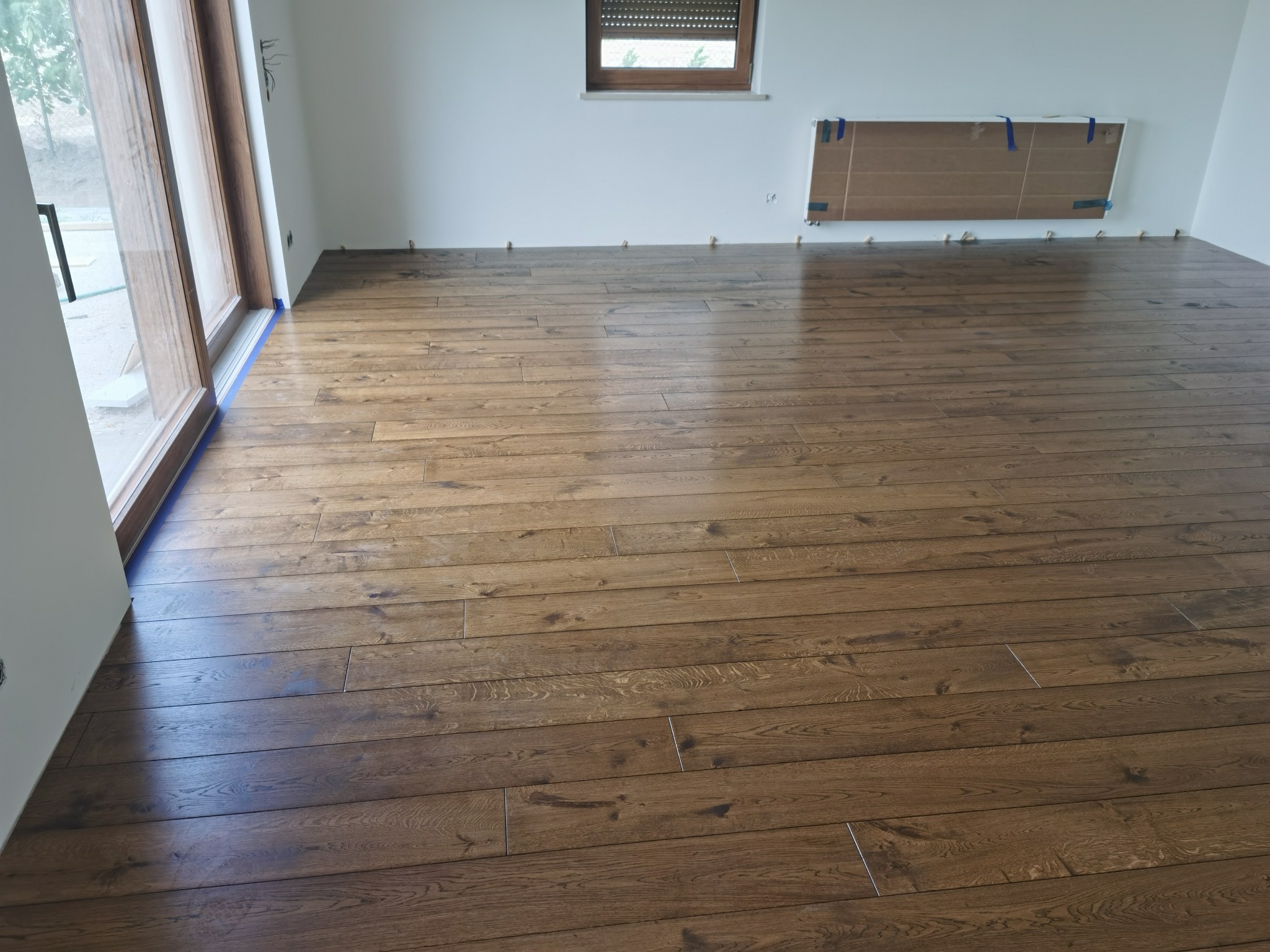 Ułożona, ciemnobrązowa podłoga drewniana z widocznymi szczelinami dylatacyjnymi przy ścianach i drzwiach balkonowych, zabezpieczonych niebieską taśmą malarską.