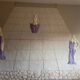 Listek Ogrody - Dekoracyjna ściana z płaskorzeźbami kobiecymi w fioletowych szatach, ułożona z kwadratowych płyt imitujących kamień, z dolną częścią wykończoną imitacją kamieni.