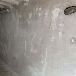 Listek Ogrody - Szara ściana z widocznymi śladami po naprawach, surowy betonowy sufit z belkami i lampa w metalowej obudowie. Wnętrze w trakcie prac wykończeniowych.
