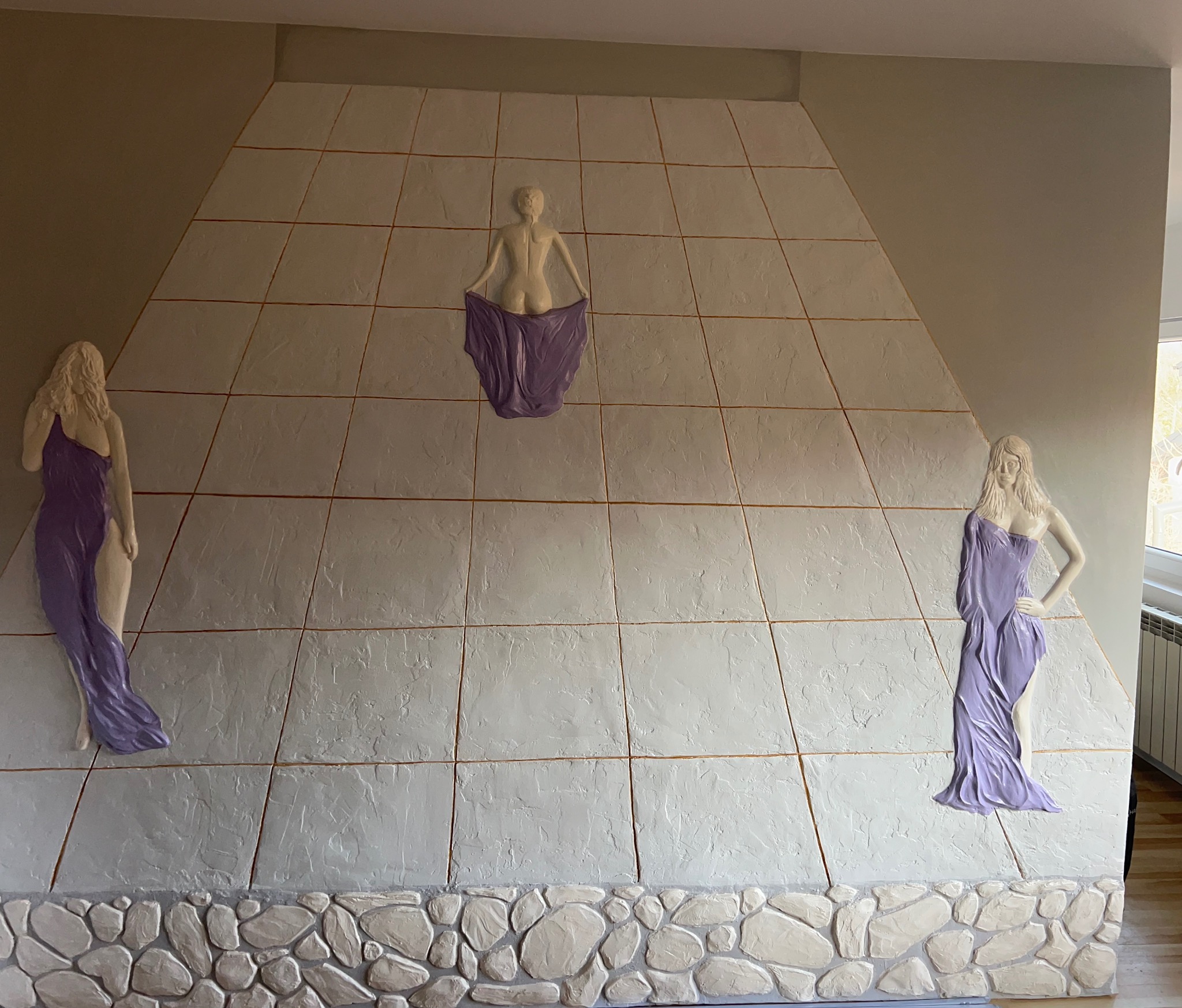 Dekoracyjna ściana z płaskorzeźbami kobiecymi w fioletowych szatach, ułożona z kwadratowych płyt imitujących kamień, z dolną częścią wykończoną imitacją kamieni.