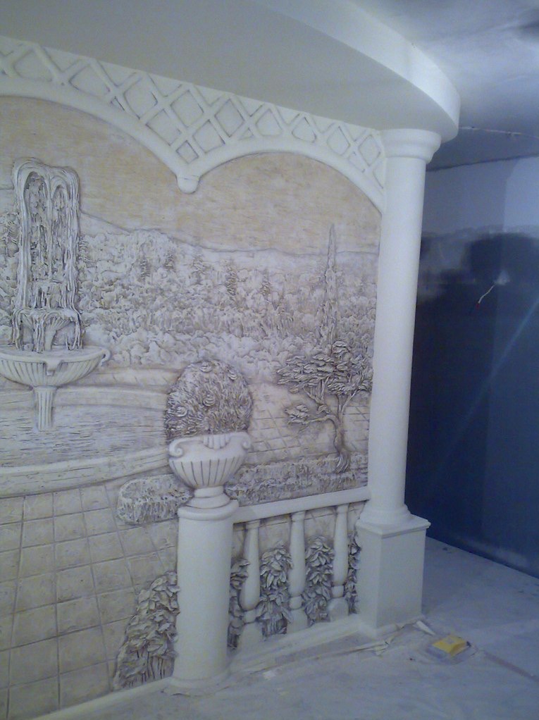 Relief ścienny z motywem ogrodu: fontanna, drzewa, balustrada z kolumną. Dekoracja w odcieniach beżu, imitująca rzeźbę. Widok z dołu, fragment pomieszczenia w tle.