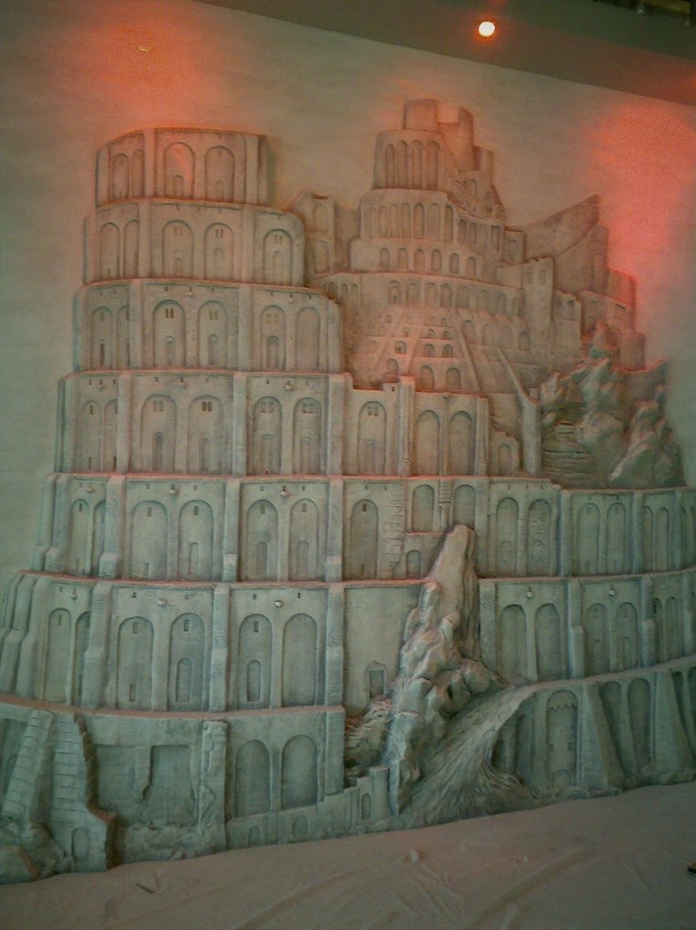 Relief na ścianie przedstawiający wielopoziomową budowlę z licznymi łukami i elementami skalnymi, oświetlony ciepłym światłem, tworzący trójwymiarowy efekt.