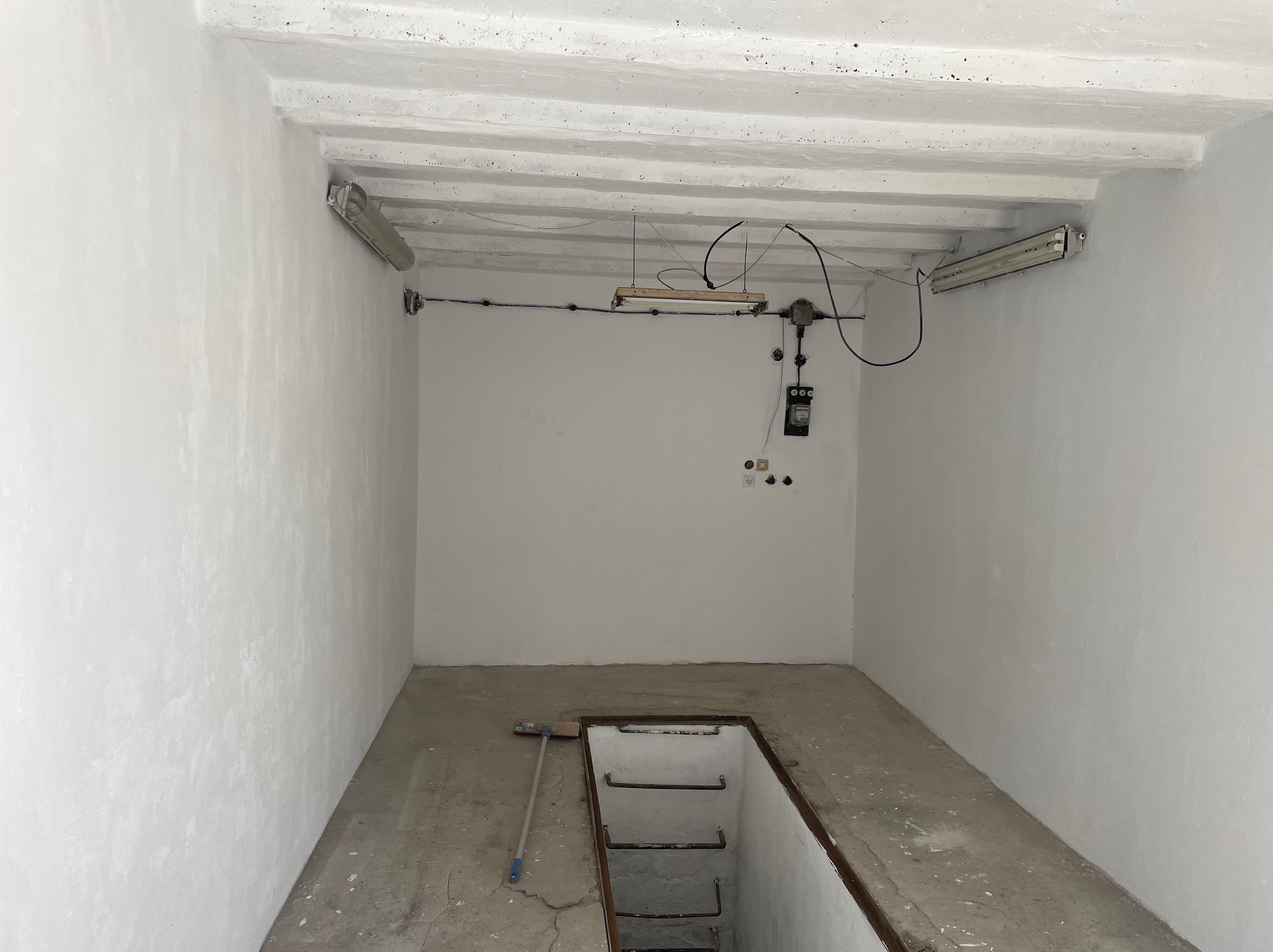 Białe, surowe wnętrze piwnicy z betonową podłogą i schodami w dół. Widoczne oświetlenie jarzeniowe i instalacja elektryczna na ścianie. Mop oparty o ścianę.