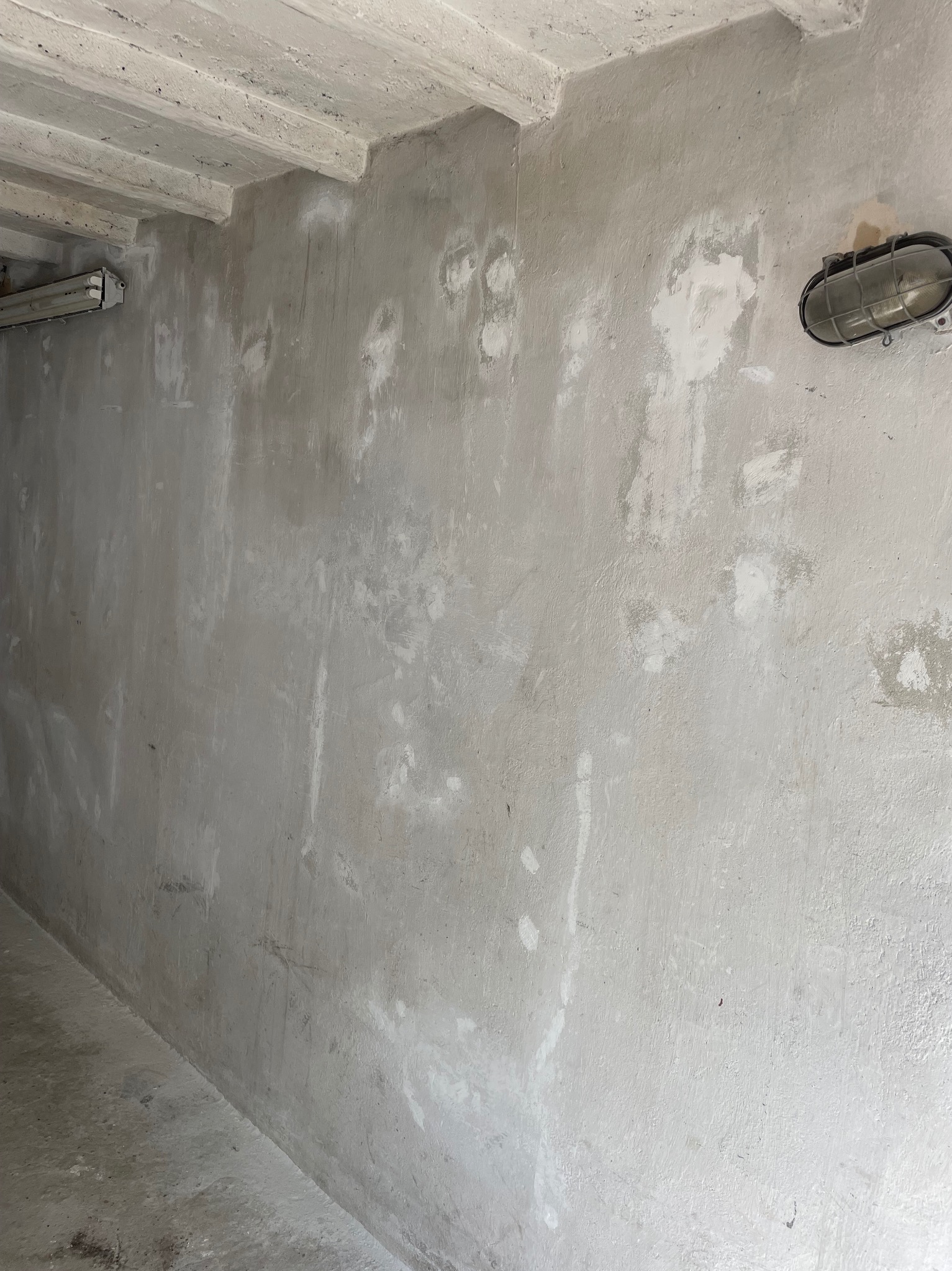 Szara ściana z widocznymi śladami po naprawach, surowy betonowy sufit z belkami i lampa w metalowej obudowie. Wnętrze w trakcie prac wykończeniowych.