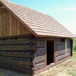 Tarasy i Elewacje z Drewna Egzotycznego , Zadaszenia - Surowy, ciemny budynek z bali z nowym dachem z dachówki ceramicznej, widoczne świeże obramowanie okna i drzwi, otoczony ziemią i trawą.