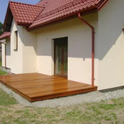 Nowoczesny taras z gładkich desek egzotycznych przylegający do jasnej elewacji domu z czerwonym dachem i rynnami, otoczony szarym żwirem i trawnikiem.