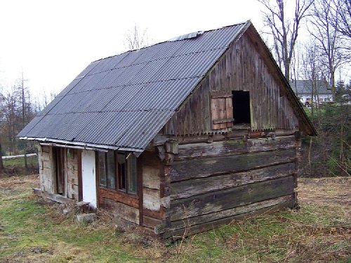 Zrujnowana, drewniana szopa z szarą, falistą blachą na dachu, z otwartym oknem na poddaszu i białą, częściowo zniszczoną stolarką okienną i drzwiową, otoczona trawą.