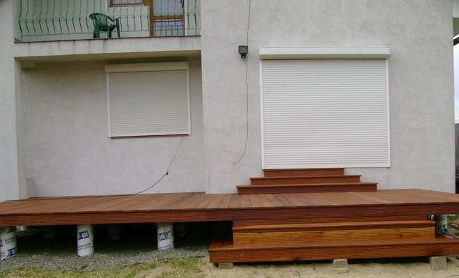 Nowoczesny taras z drewna egzotycznego przylegający do jasnej elewacji domu z roletami zewnętrznymi, widoczne podpory tarasu i schody.