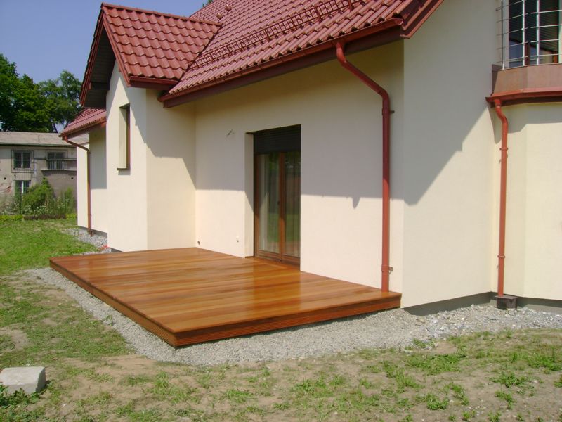 Nowoczesny taras z gładkich desek egzotycznych przylegający do jasnej elewacji domu z czerwonym dachem i rynnami, otoczony szarym żwirem i trawnikiem.