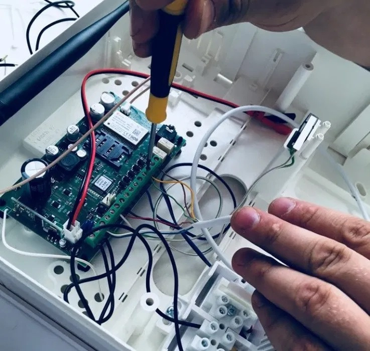 Instalacja alarmu: ręka montera przykręca śrubokrętem płytkę centrali alarmowej wewnątrz białej obudowy, widoczne liczne przewody i antena.
