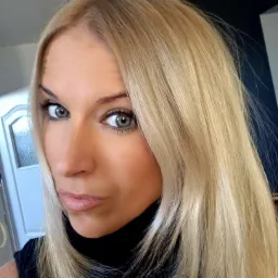 Portret kobiety z długimi, blond włosami, intensywnym spojrzeniem szaro-zielonych oczu i delikatnym makijażem.