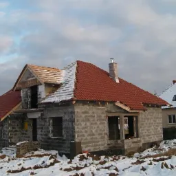 Dom w trakcie budowy w zimowej scenerii, z czerwonym dachem częściowo pokrytym śniegiem, widoczne bloczki betonowe i drewniana konstrukcja dachu.