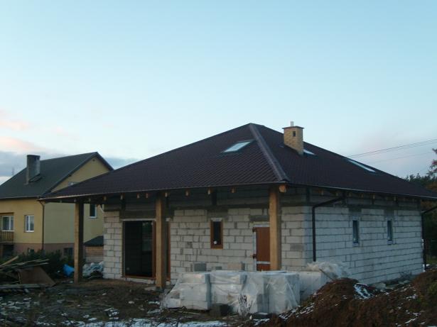 Niedokończony dom z ciemnym dachem z blachy trapezowej, widoczne okna dachowe, ściany z białych bloczków, drewniane słupy podtrzymujące okap, w tle drugi budynek.