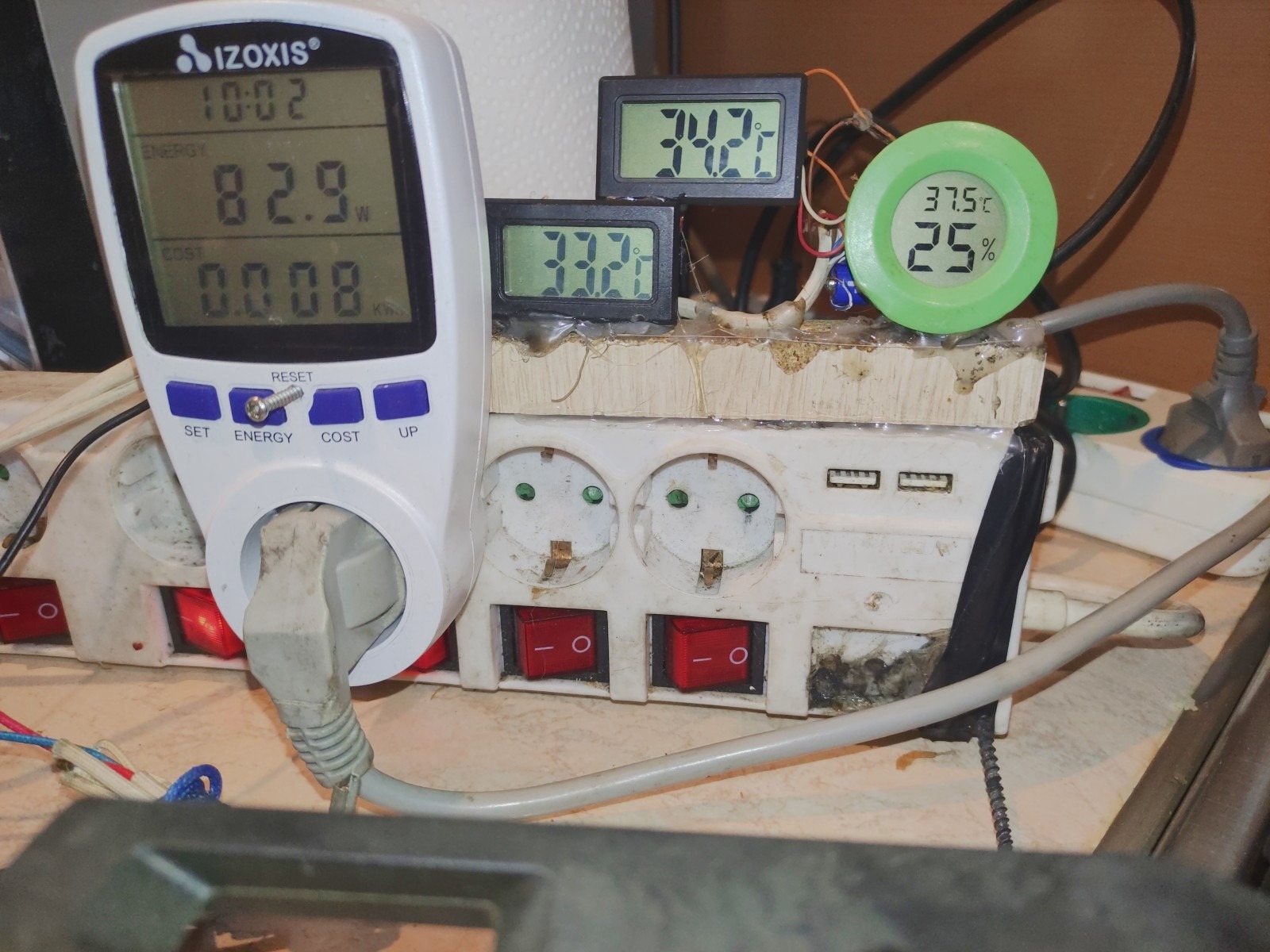 Miernik zużycia energii podłączony do listwy zasilającej, obok wyświetlacze temperatury i wilgotności, całość umieszczona na brudnej powierzchni.