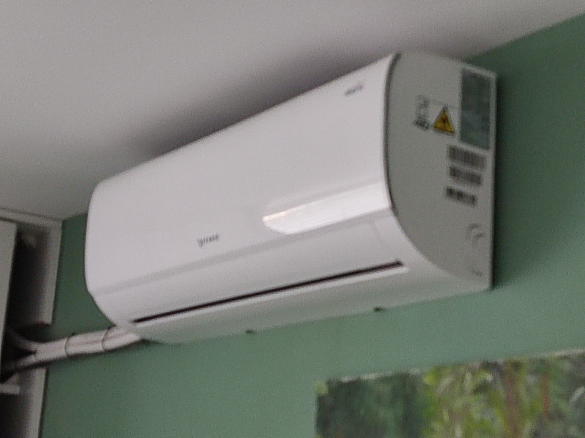 Biała jednostka wewnętrzna klimatyzacji zamontowana na zielonej ścianie, widoczne przewody i naklejka ostrzegawcza.