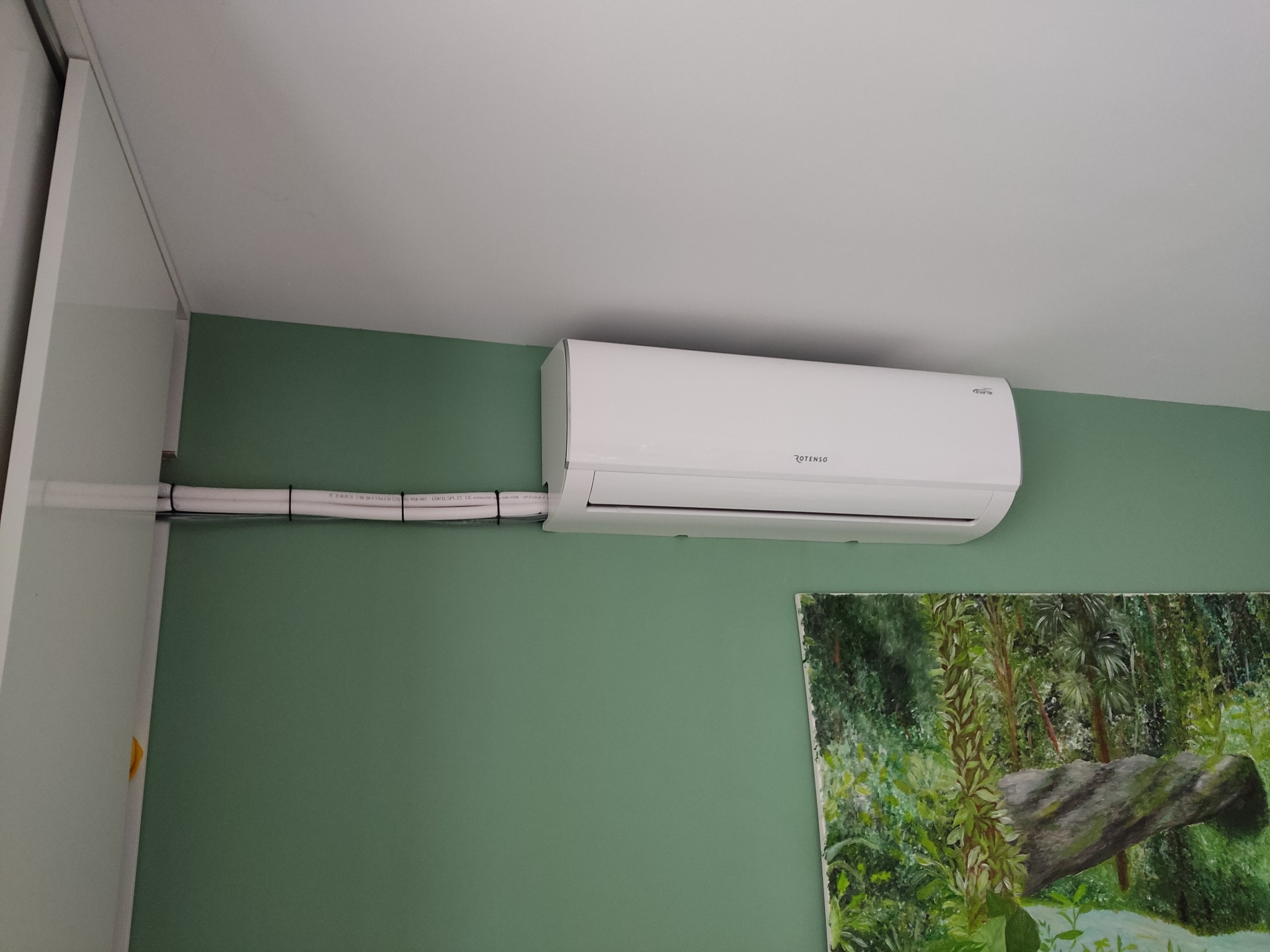 Biała jednostka klimatyzacji marki Rotenso zamontowana na zielonej ścianie z widocznymi przewodami w białej osłonie, poniżej obraz z motywem leśnym.