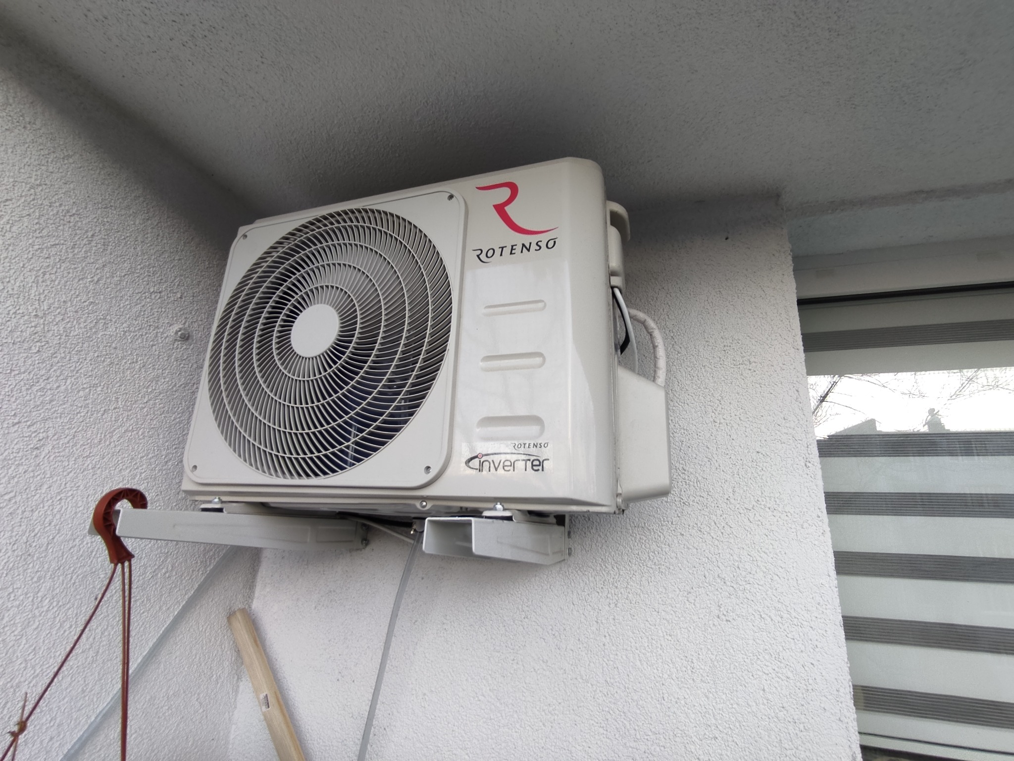 Biała jednostka zewnętrzna klimatyzacji Rotenso zamontowana na ścianie budynku z widocznym wspornikiem i przewodami.