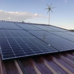 Panel słoneczny na brązowym dachu z turbiną wiatrową w tle, widok z góry, częściowo zachmurzone niebo.